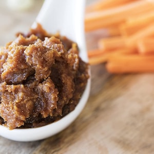 May include: A white ceramic spoon filled with a thick, brown miso paste. The spoon is sitting on a wooden surface next to a pile of orange carrot sticks.