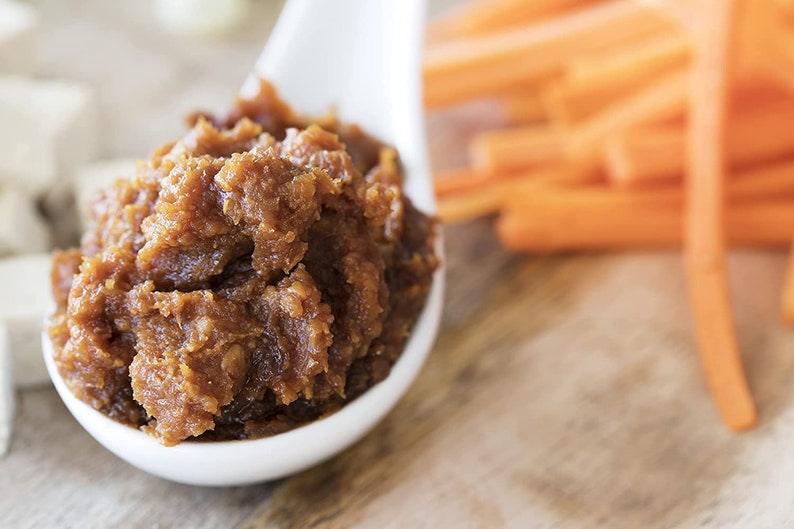 May include: A white ceramic spoon filled with a thick, brown miso paste. The spoon is sitting on a wooden surface next to a pile of orange carrot sticks.