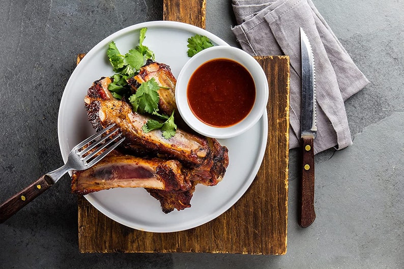 Op de afbeelding: Een wit bord met drie stukken gebarbecued varkensribben, een kleine witte kom met rode barbecuesaus en een takje koriander.