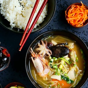 May include: A bowl of seafood soup with shrimp, octopus, mussels, and vegetables. The soup is served with a side of white rice and a small bowl of pickled vegetables.