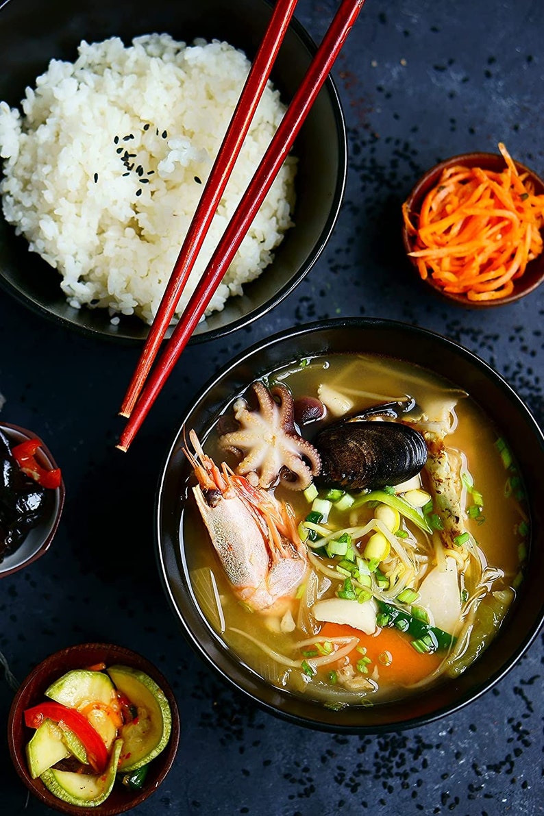 May include: A bowl of seafood soup with shrimp, octopus, mussels, and vegetables. The soup is served with a side of white rice and a small bowl of pickled vegetables.