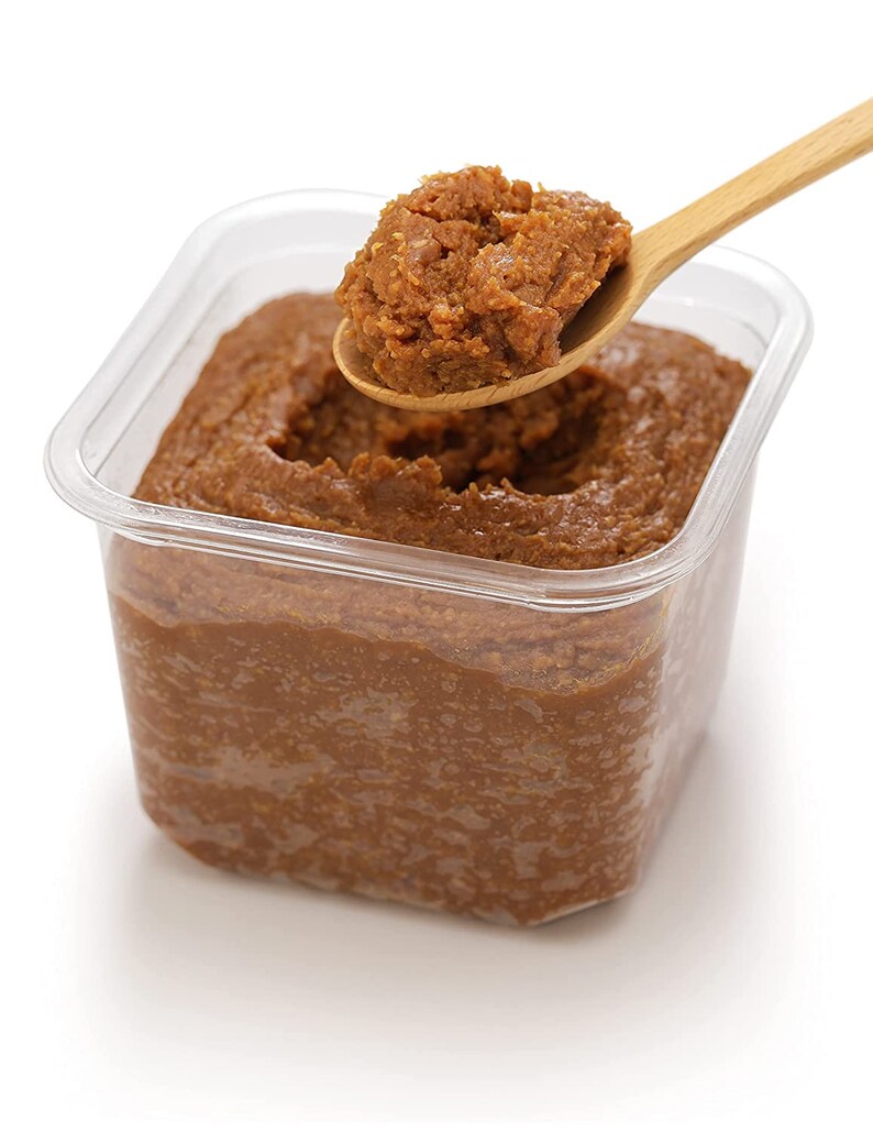 May include: A close-up of a container of brown miso paste with a spoonful of miso being lifted out of the container.