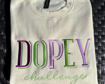 May include: Cream-coloured sweatshirt featuring the embroidered word "DOPEY" in purple and green, and the word "challenge" in green script. The sweatshirt is folded on a dark textured surface.