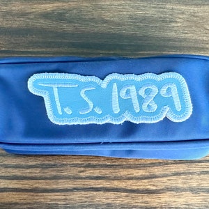 May include: A blue pencil case with a light blue embroidered patch that reads "T.S. 1989". The pencil case is rectangular with a zipper closure and a small loop on the side.