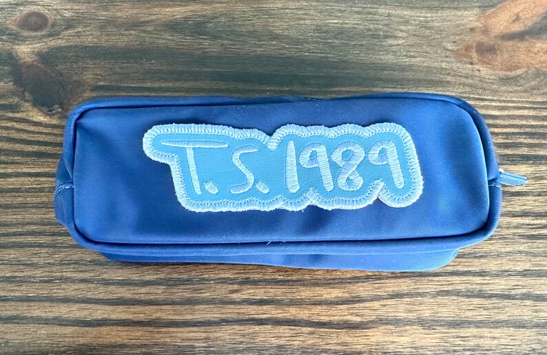 May include: A blue pencil case with a light blue embroidered patch that reads "T.S. 1989". The pencil case is rectangular with a zipper closure and a small loop on the side.