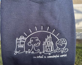Op de afbeelding: Marineblauw sweatshirt met wit geborduurd ontwerp van een kasteel, boom en bol onder een zon. De tekst "... what a wonderful world" staat eronder. Het sweatshirt wordt door een persoon omhoog gehouden.