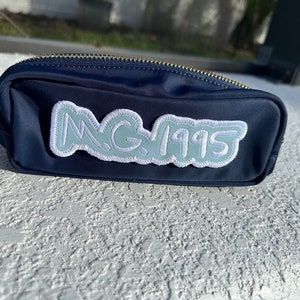 May include: Navy blue pencil case with a gold zipper. The case has a light blue patch with the text "M.G. 1995" in a cloud-like design. The case is sitting on a white textured surface.