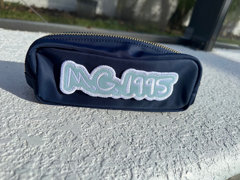 May include: Navy blue pencil case with a gold zipper. The case has a light blue patch with the text "M.G. 1995" in a cloud-like design. The case is sitting on a white textured surface.