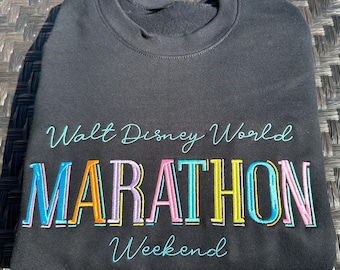 Könnte beinhalten: Dunkelgraues Sweatshirt mit dem gestickten Text "Walt Disney World Marathon Weekend" in bunten Buchstaben. Das Sweatshirt ist gefaltet und scheint aus einem weichen Material zu sein, ideal für ein Marathon-Event oder Disney-Fan.