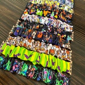 May include: A stack of colorful fabric watch bands with Halloween-themed prints, including Mickey Mouse, pumpkins, and bats. The bands are designed to fit Apple Watch.