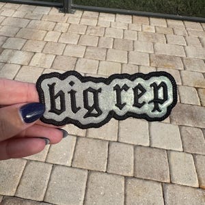 May include: A silver and black embroidered patch with the text "big rep" in a gothic font.