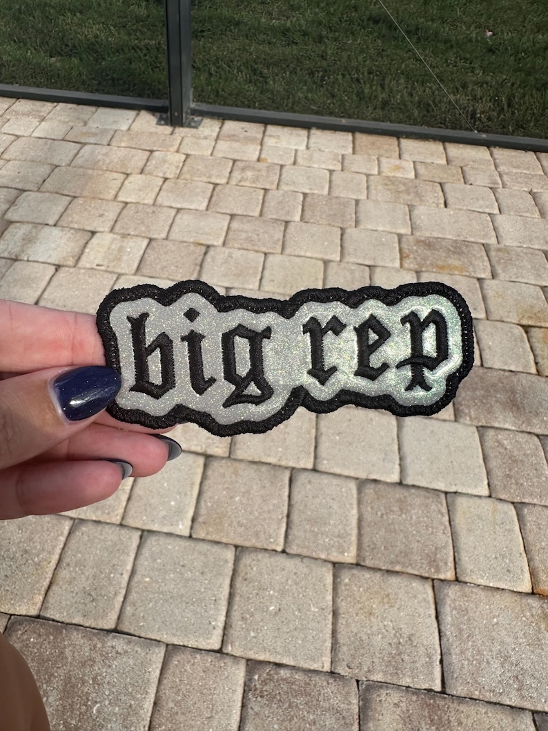 Op de afbeelding: Een zilveren en zwarte geborduurde patch met de tekst "big rep" in een gotisch lettertype.