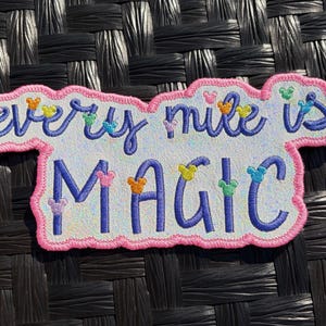 May include: Embroidered patch with the phrase "every mile is MAGIC" in blue script and block letters. The patch has a pink border and features colorful Mickey Mouse head accents.
