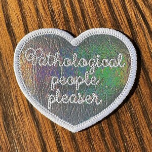 May include: A heart-shaped patch with a holographic, iridescent surface. The patch has a white embroidered border and features the words "Pathological people pleaser" in a cursive font.