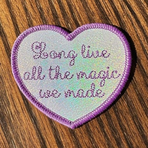 Peut inclure: Patch en forme de cœur violet avec un fond blanc et des paillettes irisées. Le patch porte le texte "Long live all the magic we made" brodé en violet.