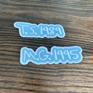 May include: Two light blue embroidered patches with white stitching. The top patch reads "T.S. 1989" and the bottom patch reads "N.G. 1995". The patches are on a wooden surface.
