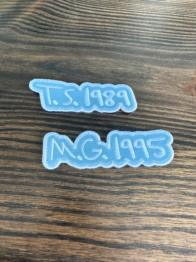 May include: Two light blue embroidered patches with white stitching. The top patch reads "T.S. 1989" and the bottom patch reads "N.G. 1995". The patches are on a wooden surface.