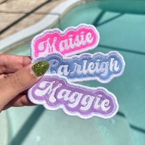 Op de afbeelding: Drie geborduurde patches met de namen Maisie, Parleigh en Maggie in witte letters op een roze, blauwe en paarse achtergrond.