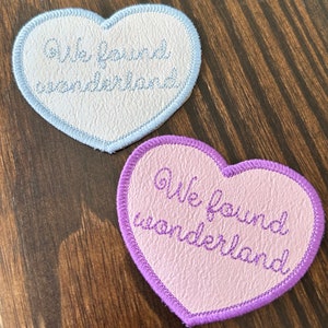 Peut inclure: Deux écussons en forme de cœur avec la broderie "We found wonderland". L'un est bleu clair avec un bord blanc, l'autre rose avec un bord violet. Les écussons sont sur une surface en bois foncé.