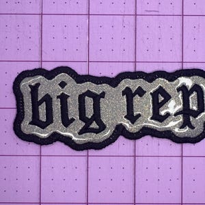 Op de afbeelding: Een zwarte en zilveren glitter strijk-op patch met de tekst "big rep" in een gotisch lettertype.