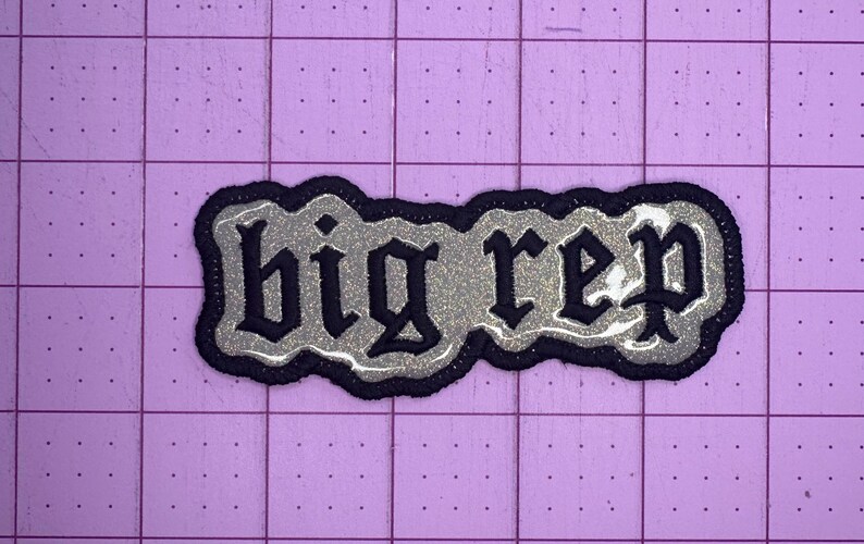 Op de afbeelding: Een zwarte en zilveren glitter strijk-op patch met de tekst "big rep" in een gotisch lettertype.