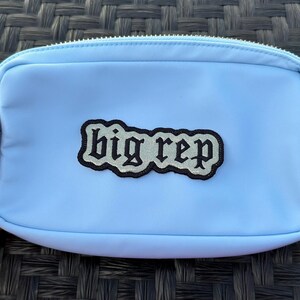 Op de afbeelding: Een lichtblauwe ritszak met een zwart-wit geborduurde patch met de tekst "big rep".