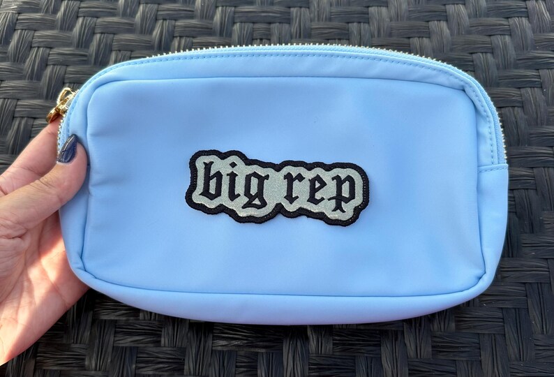 Op de afbeelding: Een lichtblauwe ritszak met een zwart-wit geborduurde patch met de tekst "big rep".
