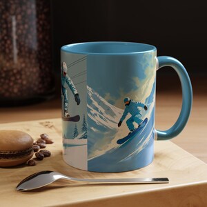 Pode incluir: Caneca de cerâmica azul com interior e alça azul claro. A caneca apresenta uma cena de esportes de inverno com snowboarders em uma montanha nevada. A caneca está em uma superfície de madeira.