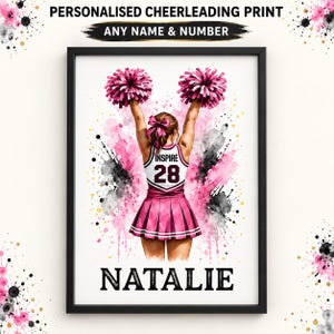 Könnte beinhalten: Ein gerahmter, personalisierter Cheerleading-Druck, der eine Cheerleaderin von hinten zeigt, die rosa Pompons hält und eine weiß-rosa Uniform mit dem Text INSPIRE und der Nummer 28 trägt, mit dem Namen NATALIE in schwarzer Schrift darunter.