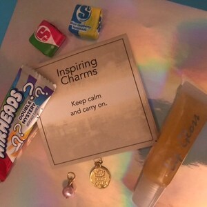 May include: A small card with the text "Inspiring Charms" and "Keep calm and carry on."  The card is surrounded by various items, including a pink and white Airheads candy bar, a small bag of Skittles, a gold charm, and a clear tube of lip gloss.