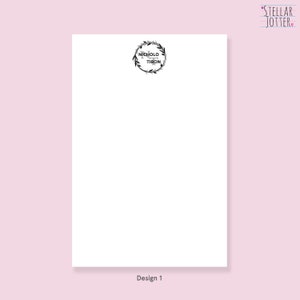 May include: A white notepad with a black wreath design and the text "Nicholo & Tibon" in the centre. The notepad has the text "Design 1" at the bottom.