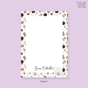 May include: A white notepad with a black and gold border featuring a repeating pattern of stars, moons, crystals, and hourglasses. The notepad has the text "June Estrella" and "Stellar Jotter" at the bottom. The notepad is labelled "Design 1".