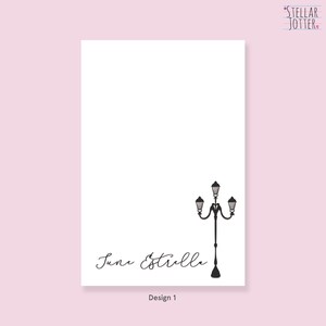 May include: A white card with the text "June Estrella" written in black cursive script. A black silhouette of a street lamp with three lights is in the top right corner. The text "Design 1" is at the bottom of the card.