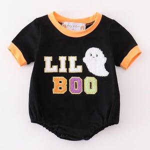 Może przedstawiać: Czarny body z krótkim rękawem i pomarańczowymi lamówkami. Body ma białego ducha z uśmiechającą się twarzą i napis "Lil Boo" kolorowymi literami.