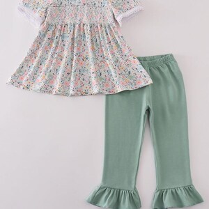 Könnte beinhalten: Ein zweiteiliges Kinder-Outfit. Das Oberteil hat einen Blumendruck, einen weißen Kragen mit orangefarbenen Kürbisstickereien und Spitzenbesatz. Die Hose ist einfarbig grün mit Rüschen am Saum.