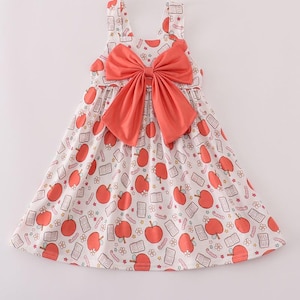 Pode incluir: Um vestido branco sem mangas com um padrão de maçãs vermelhas e livros. O vestido apresenta um grande laço coral no peito. Vestido para crianças.
