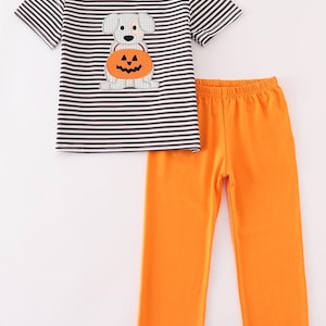 May include: A two-piece Halloween outfit for children. The top is a striped black and white short-sleeved shirt with an orange collar and a dog holding a jack-o'-lantern applique. The pants are solid orange.