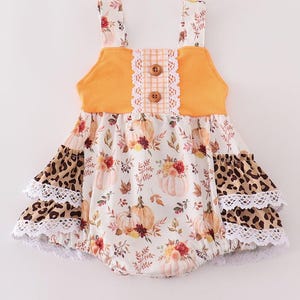Pode incluir: Um macacão de bebê branco e laranja com estampa de abóbora e flores. O macacão tem uma borda de babados e um babado de estampa de leopardo marrom na parte inferior.