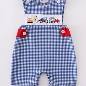 Puede incluir: Pantalón corto de bebé de cuadros azules y blancos con ribetes rojos y vehículos de construcción bordados. El pantalón corto tiene un estilo de babero con botones y un ribete rojo alrededor de las aberturas de las piernas. El bordado presenta una excavadora amarilla, un tractor azul y un camión volquete rojo.