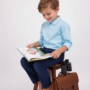 May include: A young person wearing a light blue button-down shirt and navy blue pants sits on a wooden stool, reading a book. A brown leather satchel and binoculars rest nearby. The book features colorful illustrations.