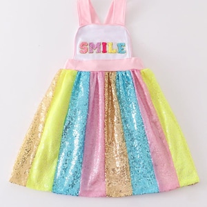 May include: A rainbow sequined dress with a pink bodice and straps. The bodice has a white patch with the word "SMILE" in colorful letters.