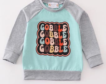 Op de afbeelding: Een lichtblauw en grijs longsleeve shirt met een kleurrijke afbeelding waarop vier keer "GOBBLE" staat.