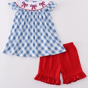 May include: A two-piece outfit featuring a blue and white gingham print top with a white collar and red bow accents. The set includes matching red shorts with ruffled hems. This outfit is suitable for children.