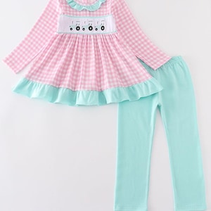 May include: A two-piece pyjama set. The top is pink and white gingham with a ruffle collar and hem. The chest has an embroidered musical note design. The trousers are solid light blue.