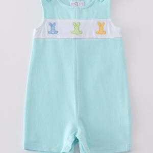 May include: A light blue romper with a white panel featuring embroidered bunnies in blue, green, and yellow. The sleeveless romper has button closures on the shoulders and at the legs. The text "Abby & Eric" is visible on the top.