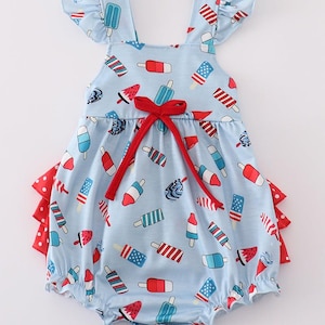 May include: Light blue baby romper with a popsicle print in red, white, and blue. The romper features a red bow and red and white polka dot ruffles. The straps have ruffled edges.