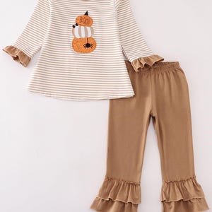 Könnte beinhalten: Ein zweiteiliges Outfit für ein Kind, bestehend aus einem langärmeligen Hemd mit weißen und braunen Streifen und einem Kürbis-Applikationen, auf dem "BOO!" steht, und einer braunen Hose mit einem doppelten Rüschen am Saum.