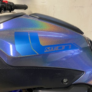 Könnte beinhalten: Nahaufnahme eines Motorrad-Benzintanks mit blauer, irisierender Lackierung. Der Tank hat ein blaues und silbernes "MT-07" Logo an der Seite.