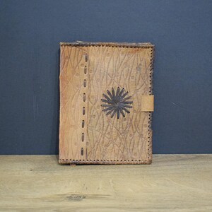 May include: A tan leather journal with a stitched border and a decorative sunburst design. The journal has a flap closure and is set against a dark blue background on a wooden surface.