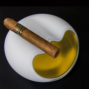 May include: White ceramic ashtray with a yellow, liquid-filled compartment. A brown cigar rests on the edge of the ashtray.